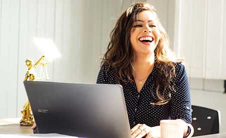 Woman wearing a dark, short-sleeve top with long, wavy hair is sitting at a desk with a silver laptop in front of her. She is laughing and looking to the right.